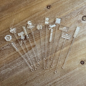 May include: A set of 10 clear acrylic drink stirrers with various designs and text, including "ONATOPP", "CAROLINA GSE", "ACHIEVER'S CLUB", "ANEA HILL", "Stoped Lion", "20", "WESTERN PR", "milago", "R", and "T".