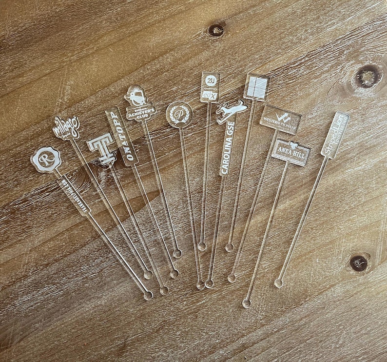 May include: A set of 10 clear acrylic drink stirrers with various designs and text, including "ONATOPP", "CAROLINA GSE", "ACHIEVER'S CLUB", "ANEA HILL", "Stoped Lion", "20", "WESTERN PR", "milago", "R", and "T".