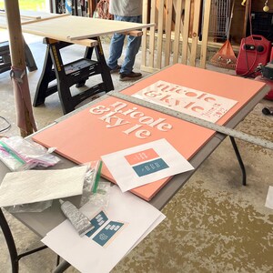 K&ouml;nnte beinhalten: Ein korallfarbenes Schild mit den Namen "nicole & kyle" in Wei&szlig; und dem Datum "June 18, 2022". Das Schild liegt auf einem Tisch mit Werkzeugen und gedruckten Designvorlagen. Die Szene deutet auf ein DIY-Hochzeitsprojekt hin.
