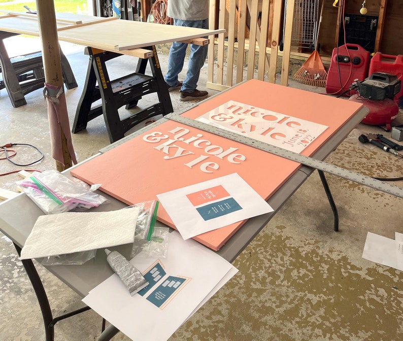 K&ouml;nnte beinhalten: Ein korallfarbenes Schild mit den Namen "nicole & kyle" in Wei&szlig; und dem Datum "June 18, 2022". Das Schild liegt auf einem Tisch mit Werkzeugen und gedruckten Designvorlagen. Die Szene deutet auf ein DIY-Hochzeitsprojekt hin.