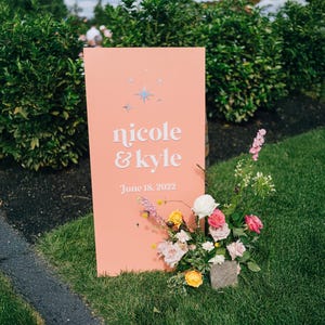 K&ouml;nnte beinhalten: Ein korallfarbenes Hochzeitsschild mit den Namen "nicole & kyle" in wei&szlig;er Schrift und dem Datum "June 18, 2022". Das Schild ist mit Sternakzenten und einem Blumenarrangement aus rosa, gelben und wei&szlig;en Blumen verziert.