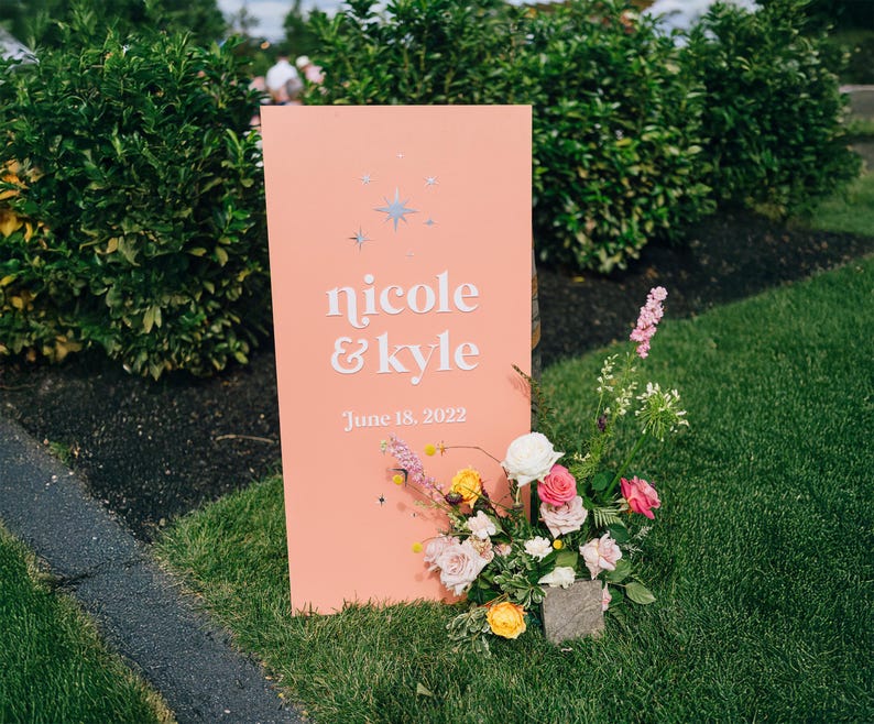 K&ouml;nnte beinhalten: Ein korallfarbenes Hochzeitsschild mit den Namen "nicole & kyle" in wei&szlig;er Schrift und dem Datum "June 18, 2022". Das Schild ist mit Sternakzenten und einem Blumenarrangement aus rosa, gelben und wei&szlig;en Blumen verziert.