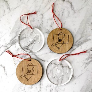 Puede incluir: Cuatro adornos navide&ntilde;os, dos son de acr&iacute;lico transparente y dos de madera. Cada adorno presenta un contorno blanco del estado de Nueva Jersey con un coraz&oacute;n en el centro. Los adornos cuelgan de cuerdas rojas y doradas.