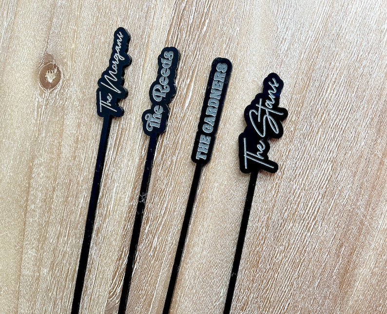 May include: Four black acrylic cocktail stirrers with silver lettering. The stirrers are labeled "The Morgans", "The Reeds", "The Gardners", and "The Stans".