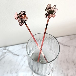 K&ouml;nnte beinhalten: Zwei ros&eacute;goldene Cocktailr&uuml;hrer mit personalisiertem Text. Ein R&uuml;hrer sagt "Mama Brie" mit einem Herzsymbol. Der andere R&uuml;hrer sagt "Baby Ezra".