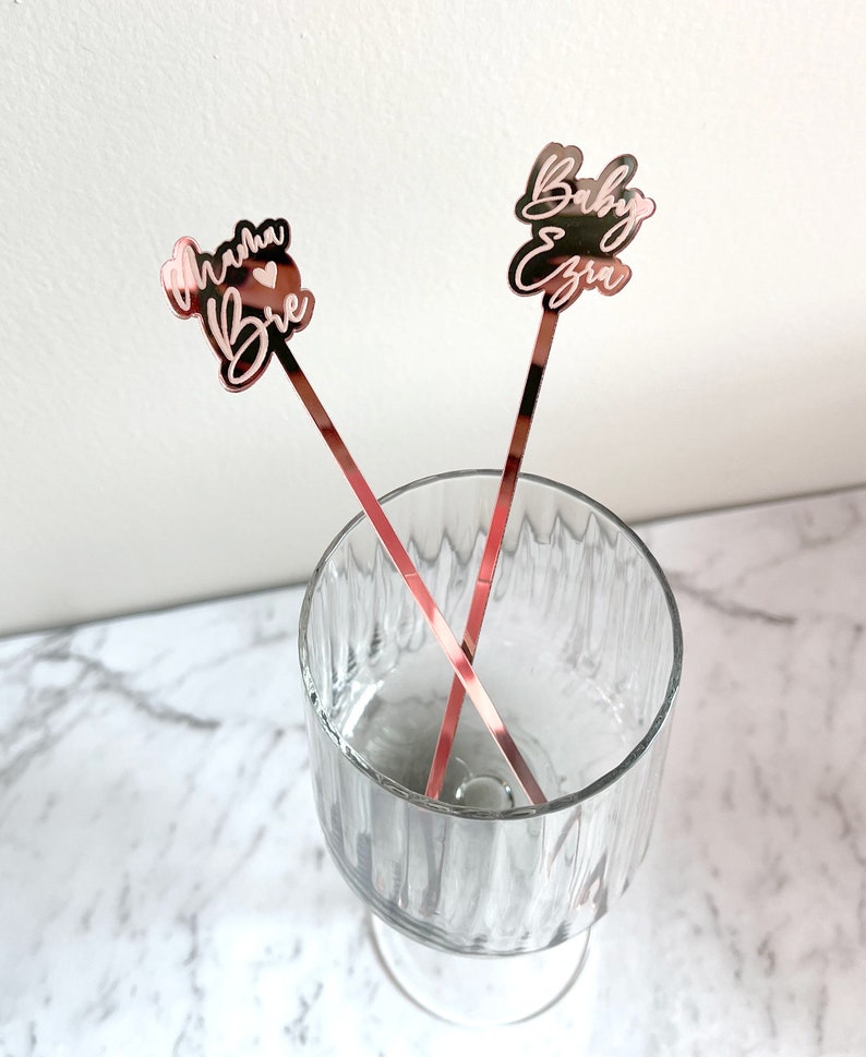 K&ouml;nnte beinhalten: Zwei ros&eacute;goldene Cocktailr&uuml;hrer mit personalisiertem Text. Ein R&uuml;hrer sagt "Mama Brie" mit einem Herzsymbol. Der andere R&uuml;hrer sagt "Baby Ezra".