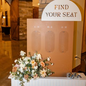 K&ouml;nnte beinhalten: Ein Hochzeits-Sitzplan in Pfirsichfarbe mit den Worten "FIND YOUR SEAT" in goldfarbenen Buchstaben. Der Plan hat drei transparente Tafeln f&uuml;r die Namen der G&auml;ste. Darunter befindet sich eine wei&szlig;e Bank mit einem Blumenarrangement.