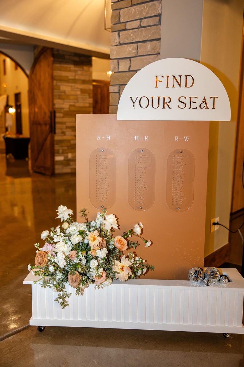 K&ouml;nnte beinhalten: Ein Hochzeits-Sitzplan in Pfirsichfarbe mit den Worten "FIND YOUR SEAT" in goldfarbenen Buchstaben. Der Plan hat drei transparente Tafeln f&uuml;r die Namen der G&auml;ste. Darunter befindet sich eine wei&szlig;e Bank mit einem Blumenarrangement.
