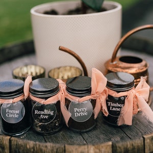 May include: Four small glass jars with black lids, each labeled with a different street name: Forest Circle, Reading Ave, Perry Drive, and Timber Lane. The jars are tied with peach-colored ribbons. A copper watering can and a potted plant are in the background.