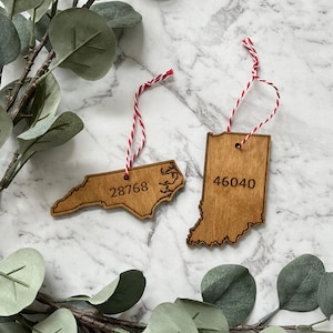 May include: Two wooden ornaments shaped like the states of North Carolina and Indiana. The North Carolina ornament has the zip code 28768 engraved on it, and the Indiana ornament has the zip code 46040 engraved on it.