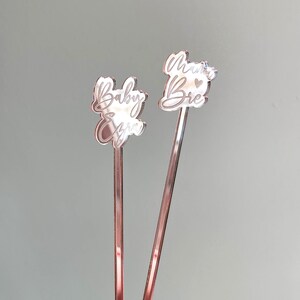 K&ouml;nnte beinhalten: Zwei ros&eacute;farbene R&uuml;hrst&auml;bchen mit graviertem Text. Ein St&auml;bchen sagt "Baby Girl" und das andere sagt "Mama Bear" mit einem kleinen Herzen.