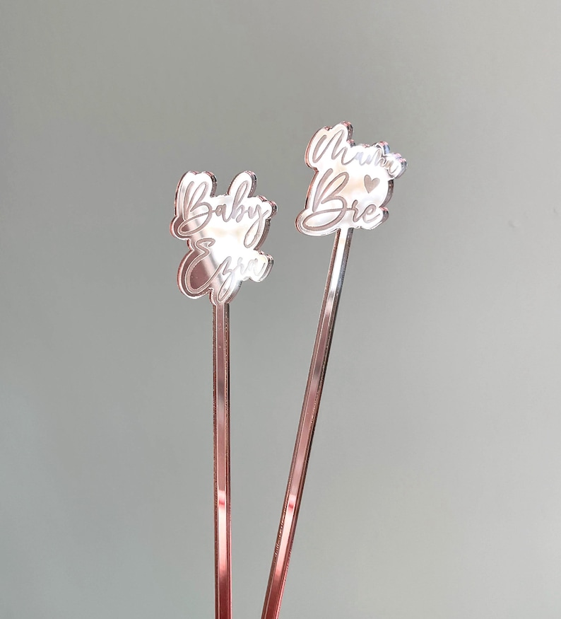 K&ouml;nnte beinhalten: Zwei ros&eacute;farbene R&uuml;hrst&auml;bchen mit graviertem Text. Ein St&auml;bchen sagt "Baby Girl" und das andere sagt "Mama Bear" mit einem kleinen Herzen.