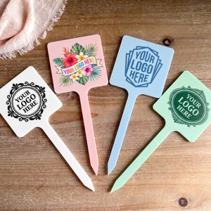 May include: Four square plant markers with pointed stakes in white, pink, light blue, and light green. Each marker features a unique design and the text "YOUR LOGO HERE" in black or blue ink, including floral patterns and decorative circular frames.