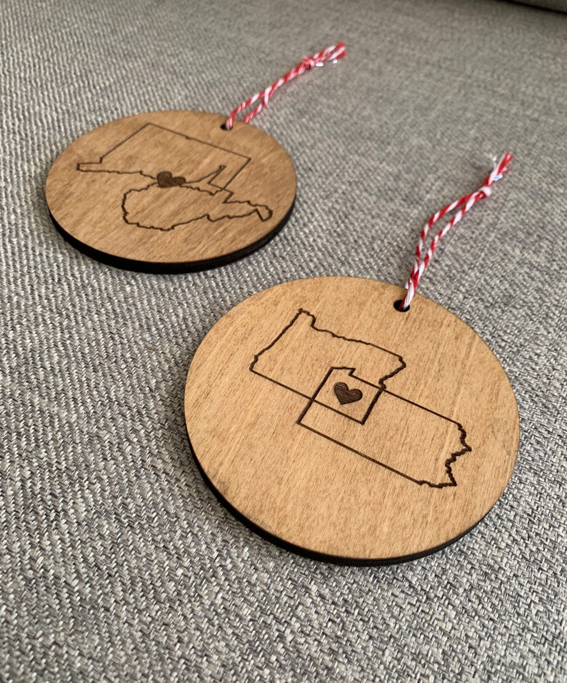 Puede incluir: Dos adornos de madera con forma de los estados de Pensilvania y Ohio con un coraz&oacute;n en el medio de Ohio. Los adornos cuelgan de un cordel rojo y blanco.