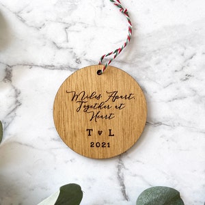 Puede incluir: Un adorno redondo de madera con el texto "Miles Apart, Together at Heart" y las iniciales "T &hearts; L" y el a&ntilde;o "2021".