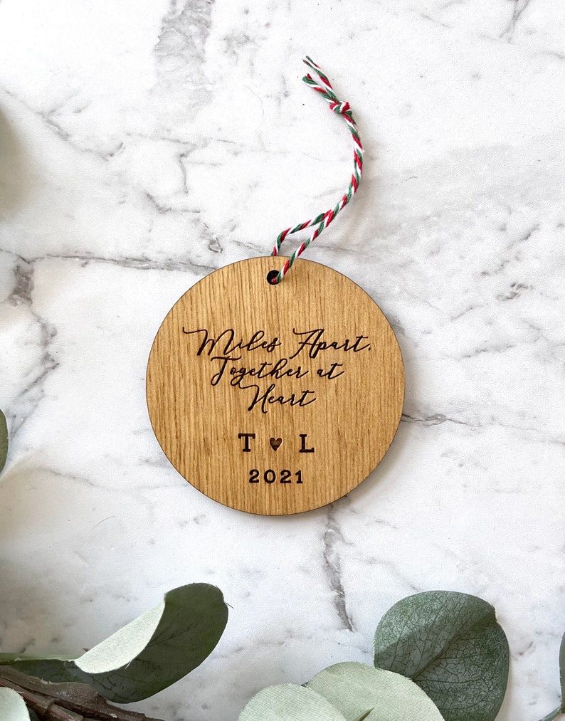 Puede incluir: Un adorno redondo de madera con el texto "Miles Apart, Together at Heart" y las iniciales "T &hearts; L" y el a&ntilde;o "2021".