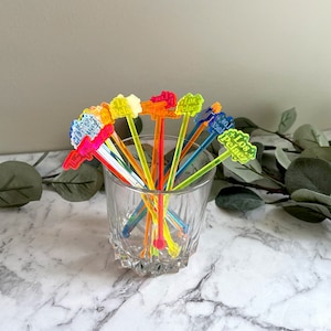 Puede incluir: Un conjunto de palillos de c&oacute;ctel de pl&aacute;stico de colores con las palabras "Los Pelotes" impresas en ellos. Los palillos est&aacute;n dispuestos en un vaso sobre una superficie de m&aacute;rmol blanco.