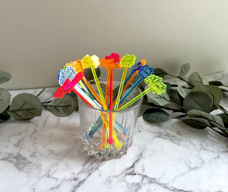 Puede incluir: Un conjunto de palillos de c&oacute;ctel de pl&aacute;stico de colores con las palabras "Los Pelotes" impresas en ellos. Los palillos est&aacute;n dispuestos en un vaso sobre una superficie de m&aacute;rmol blanco.