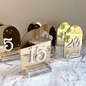 K&ouml;nnte beinhalten: Tischkarten aus goldfarbenem, verspiegeltem Acryl mit erhabenen wei&szlig;en Zahlen. Jede Zahl steht auf einem Sockel aus klarem Acryl. Die Zahlen haben eine bogenf&ouml;rmige Form, wobei die Zahlen 5, 15 und 20 sichtbar sind.