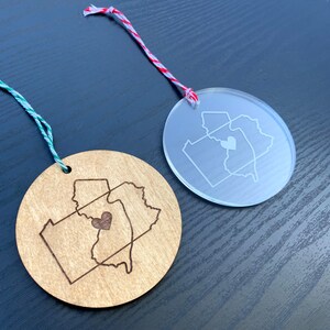 Puede incluir: Dos adornos redondos, uno de madera y otro de acr&iacute;lico transparente, ambos con un contorno de los estados de Pensilvania y Nueva Jersey con un coraz&oacute;n en el medio. El adorno de madera tiene un acabado marr&oacute;n y el adorno de acr&iacute;lico es transparente.