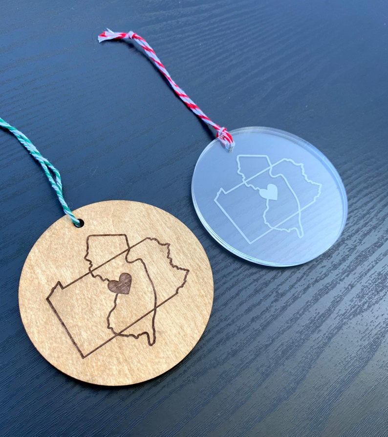 Puede incluir: Dos adornos redondos, uno de madera y otro de acr&iacute;lico transparente, ambos con un contorno de los estados de Pensilvania y Nueva Jersey con un coraz&oacute;n en el medio. El adorno de madera tiene un acabado marr&oacute;n y el adorno de acr&iacute;lico es transparente.