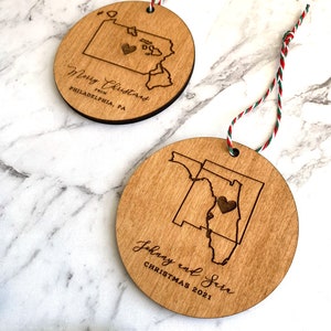 Puede incluir: Dos adornos navide&ntilde;os de madera con forma de los estados de Pensilvania y Florida. El adorno de Pensilvania tiene el texto "Merry Christmas from Philadelphia, PA" y un peque&ntilde;o coraz&oacute;n en el centro del estado. El adorno de Florida tiene el texto "Johnny and Sara Christmas 2021" y un peque&ntilde;o coraz&oacute;n en el centro del estado.