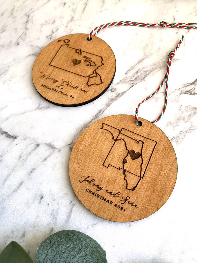 Puede incluir: Dos adornos navide&ntilde;os de madera con forma de los estados de Pensilvania y Florida. El adorno de Pensilvania tiene el texto "Merry Christmas from Philadelphia, PA" y un peque&ntilde;o coraz&oacute;n en el centro del estado. El adorno de Florida tiene el texto "Johnny and Sara Christmas 2021" y un peque&ntilde;o coraz&oacute;n en el centro del estado.