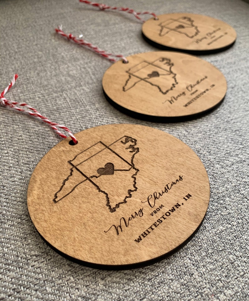 Puede incluir: Tres adornos de madera con forma del estado de Indiana con un coraz&oacute;n en el centro. Cada adorno tiene el texto "Merry Christmas from Whitestown, IN" grabado en &eacute;l.