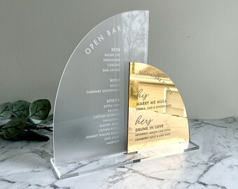 Könnte beinhalten: Ein klares Acrylschild mit einem daran befestigten goldenen Spiegelschild. Das Acrylschild listet eine Vielzahl von Bier, Wein und Spirituosen auf. Das goldene Spiegelschild listet zwei Signature-Drinks auf: "Marry Me Mula" und "Drunk in Love".
