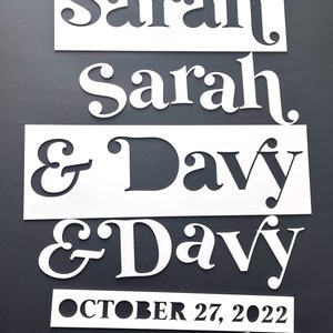 K&ouml;nnte beinhalten: Wei&szlig;e, lasergeschnittene Holzschilder mit den Namen "Sarah" und "Davy" und dem Datum "27. Oktober 2022". Die Namen und das Datum sind in einer dekorativen Schriftart dargestellt. Die Schilder befinden sich auf einem dunkelgrauen Hintergrund.