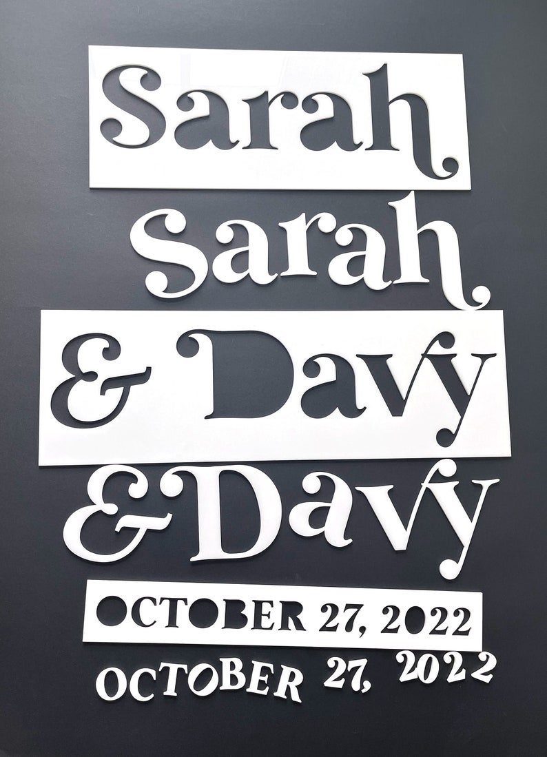K&ouml;nnte beinhalten: Wei&szlig;e, lasergeschnittene Holzschilder mit den Namen "Sarah" und "Davy" und dem Datum "27. Oktober 2022". Die Namen und das Datum sind in einer dekorativen Schriftart dargestellt. Die Schilder befinden sich auf einem dunkelgrauen Hintergrund.