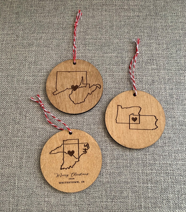 Puede incluir: Tres adornos navide&ntilde;os de madera con los contornos de los estados de Virginia Occidental, Pensilvania e Indiana. Cada adorno tiene un coraz&oacute;n en la ubicaci&oacute;n de la ciudad de origen. El adorno de Indiana tambi&eacute;n incluye el texto "Merry Christmas from Whitestown, IN".