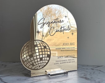 May include: Gold mirror sign with a disco ball design and the text "Signature Cocktails". The sign also lists two cocktail recipes: "Jojo Juice" and "Spicy Santiago".