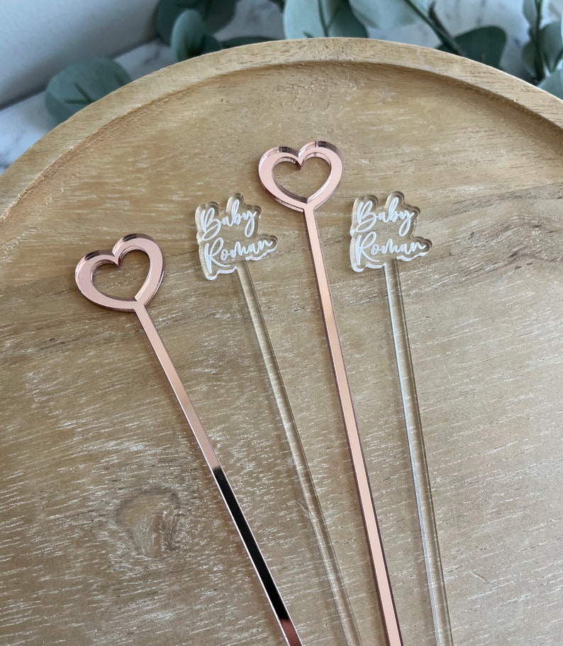 K&ouml;nnte beinhalten: Drei ros&eacute;goldene Acryl-Getr&auml;nkr&uuml;hrer mit herzf&ouml;rmigen Aufs&auml;tzen. Jeder R&uuml;hrer tr&auml;gt den Text "Baby Rohan" eingraviert.