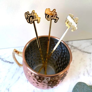 K&ouml;nnte beinhalten: Drei goldfarbene Cocktailr&uuml;hrer aus Metall mit den personalisierten Namen "Randy & Becky", die oben eingraviert sind. Die R&uuml;hrer befinden sich in einem Kupfer-Moscow-Mule-Becher.