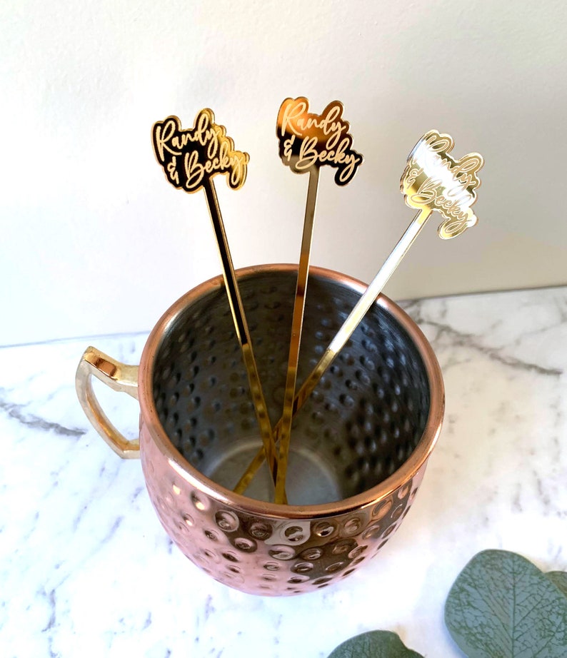 K&ouml;nnte beinhalten: Drei goldfarbene Cocktailr&uuml;hrer aus Metall mit den personalisierten Namen "Randy & Becky", die oben eingraviert sind. Die R&uuml;hrer befinden sich in einem Kupfer-Moscow-Mule-Becher.