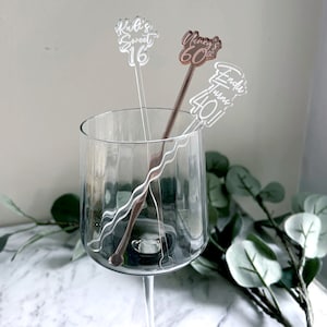 Puede incluir: Tres agitadores de bebidas de acr&iacute;lico transparente con texto personalizado. Los agitadores est&aacute;n en una copa de vino transparente. El texto en los agitadores dice "Kail's Sweet 16", "Nancy's 60th" y "Linda's Turning 40".