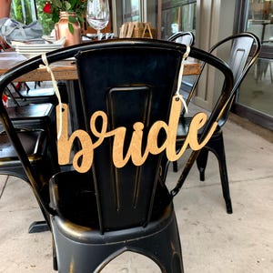 K&ouml;nnte beinhalten: Ein Holzschild mit dem Wort "Braut" in Schreibschrift h&auml;ngt an der R&uuml;ckseite eines schwarzen Metallstuhls. Der Stuhl geh&ouml;rt zu einer Reihe von St&uuml;hlen um einen Tisch, der f&uuml;r eine Feier gedeckt ist. Ein Strau&szlig; roter und wei&szlig;er Blumen ist im Hintergrund zu sehen.