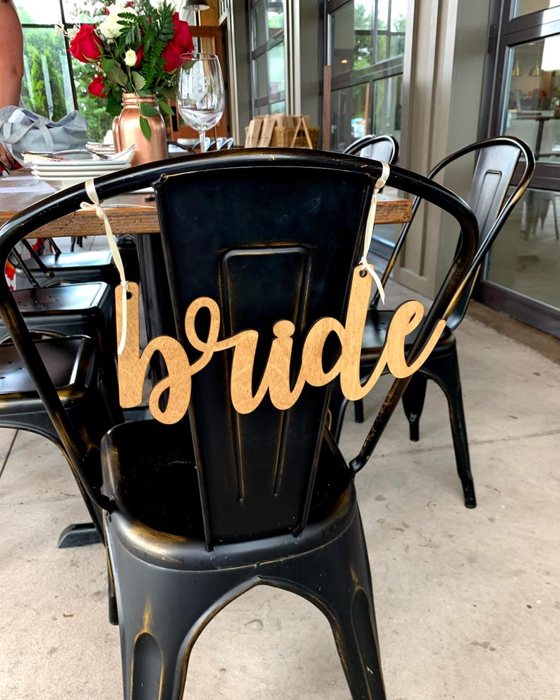 K&ouml;nnte beinhalten: Ein Holzschild mit dem Wort "Braut" in Schreibschrift h&auml;ngt an der R&uuml;ckseite eines schwarzen Metallstuhls. Der Stuhl geh&ouml;rt zu einer Reihe von St&uuml;hlen um einen Tisch, der f&uuml;r eine Feier gedeckt ist. Ein Strau&szlig; roter und wei&szlig;er Blumen ist im Hintergrund zu sehen.