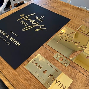 K&ouml;nnte beinhalten: Ein schwarzes Schild mit goldfarbenen Buchstaben, auf dem "It was always you" und die Namen "Sam & Kevin" mit dem Datum "11.11.22" stehen. Goldene Spiegelausschnitte des gleichen Textes sind ebenfalls vorhanden.