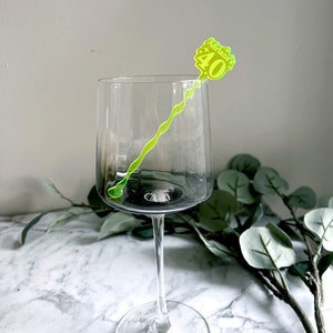 Puede incluir: Una copa de vino de vidrio transparente con un agitador de bebidas de pl&aacute;stico amarillo y verde. El agitador tiene el texto "Adriane's 40".