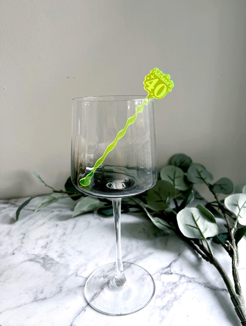 Puede incluir: Una copa de vino de vidrio transparente con un agitador de bebidas de pl&aacute;stico amarillo y verde. El agitador tiene el texto "Adriane's 40".