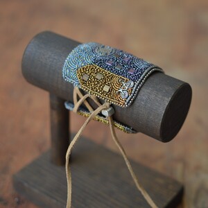 May include: A beaded bracelet with a brown leather cord and a silver metal clasp. The bracelet is displayed on a wooden stand.