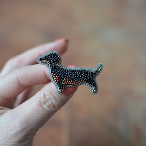 Puede incluir: Un broche de perro salchicha con cuentas. El perro es negro con detalles en marrón y dorado, y un contorno gris. El broche está hecho de pequeñas cuentas y tiene un diseño detallado. El perro está de pie.