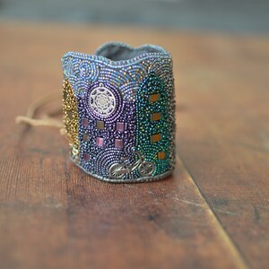 May include: A beaded bracelet with a colorful design featuring a bicycle, geometric shapes, and a circular pattern. The bracelet is made of small, iridescent beads in shades of gold, purple, green, and blue.