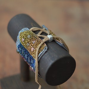 May include: A beaded bracelet with a brown leather strap and a silver metal accent. The bracelet is made with iridescent blue, gold, and copper beads.