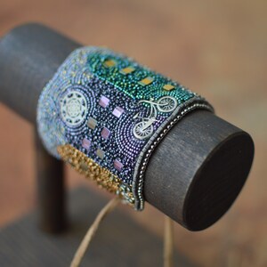 May include: A beaded bracelet with a colorful design featuring a bicycle, squares, and circles. The bracelet is displayed on a wooden stand.