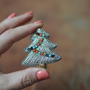 Puede incluir: Un broche de árbol de Navidad plateado con diamantes de imitación naranja, azul y morado. El broche se sostiene en una mano con esmalte de uñas rosa.