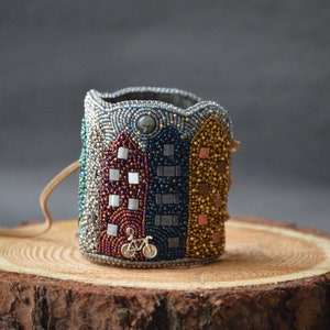 May include: A beaded bracelet with a cityscape design featuring colorful buildings and a small bicycle. The bracelet is made with a variety of beads in shades of blue, green, gold, and red.