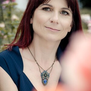 Puede incluir: Una mujer con el pelo largo y rojo que lleva una blusa azul oscuro sin mangas con un collar de cuentas con un colgante de piedra azul.